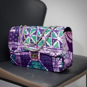 Yelé Print Embellished Maxi Bag resting on a black leather chair showcasing the bold purple and teal cultural Unity print and structured shape.